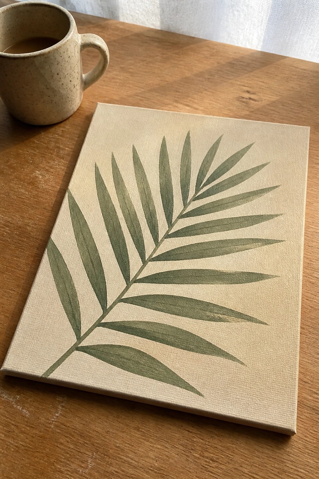 Green palm frond painting on beige canvas beside speckled mug on wood table