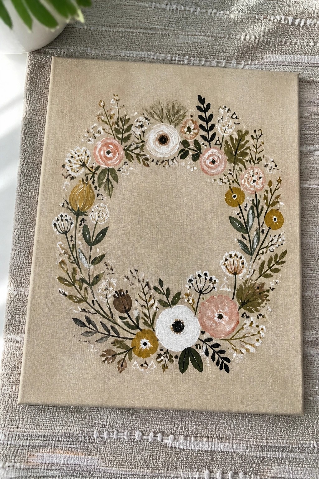 A canvas painting of a circular floral wreath with pink, white, yellow, and green flowers on a beige background.