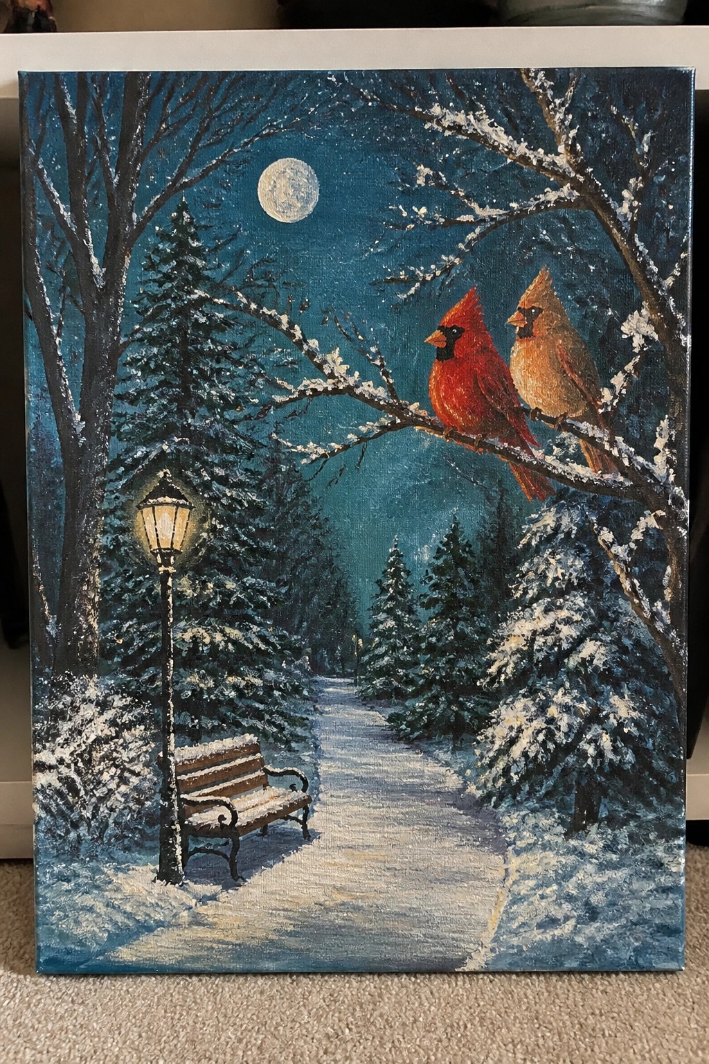 Winter painting of two cardinals on snowy branch under full moon with glowing lamppost and bench