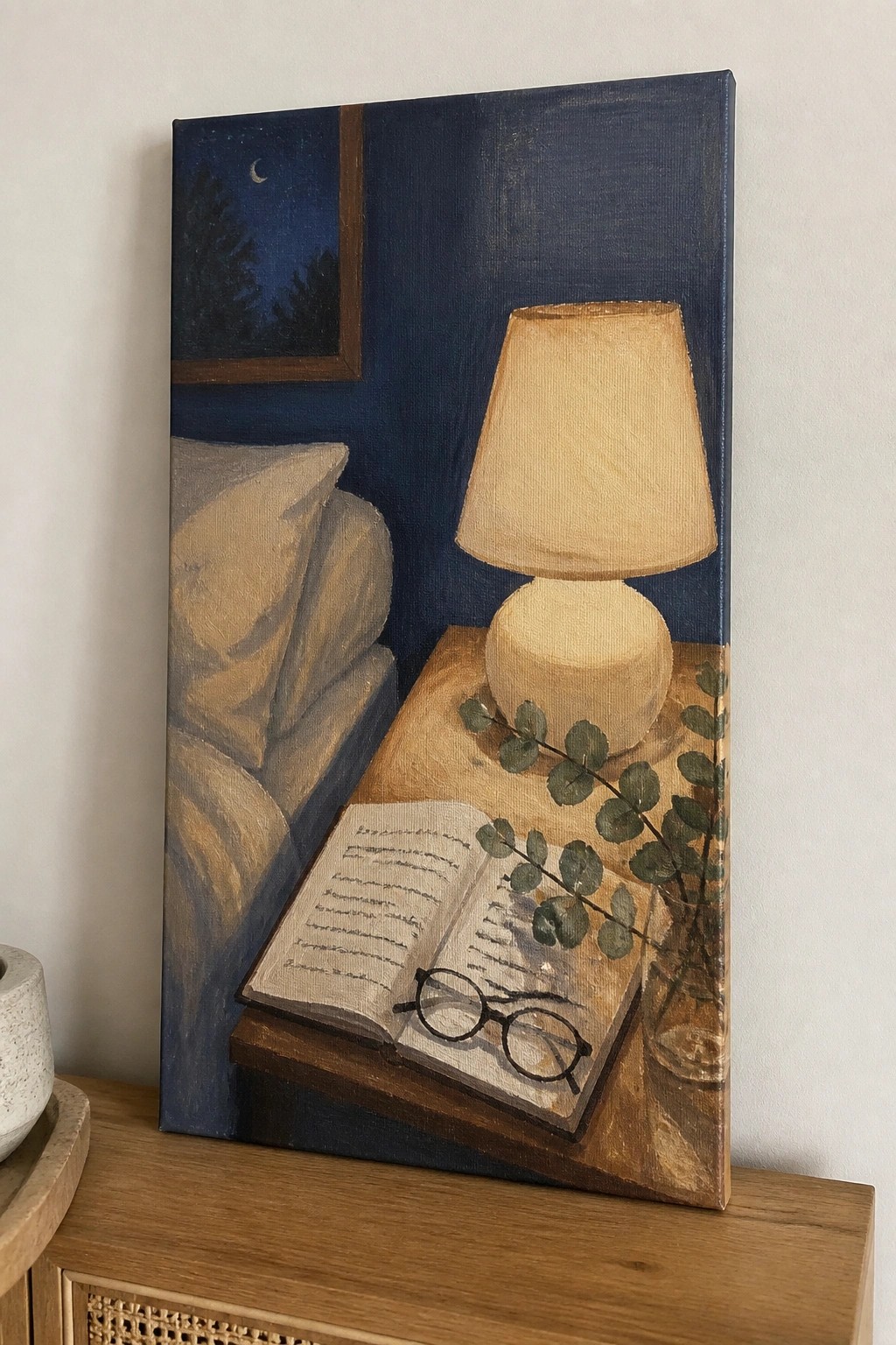 Painting of open book with glasses beside glowing lamp and eucalyptus on nightstand.