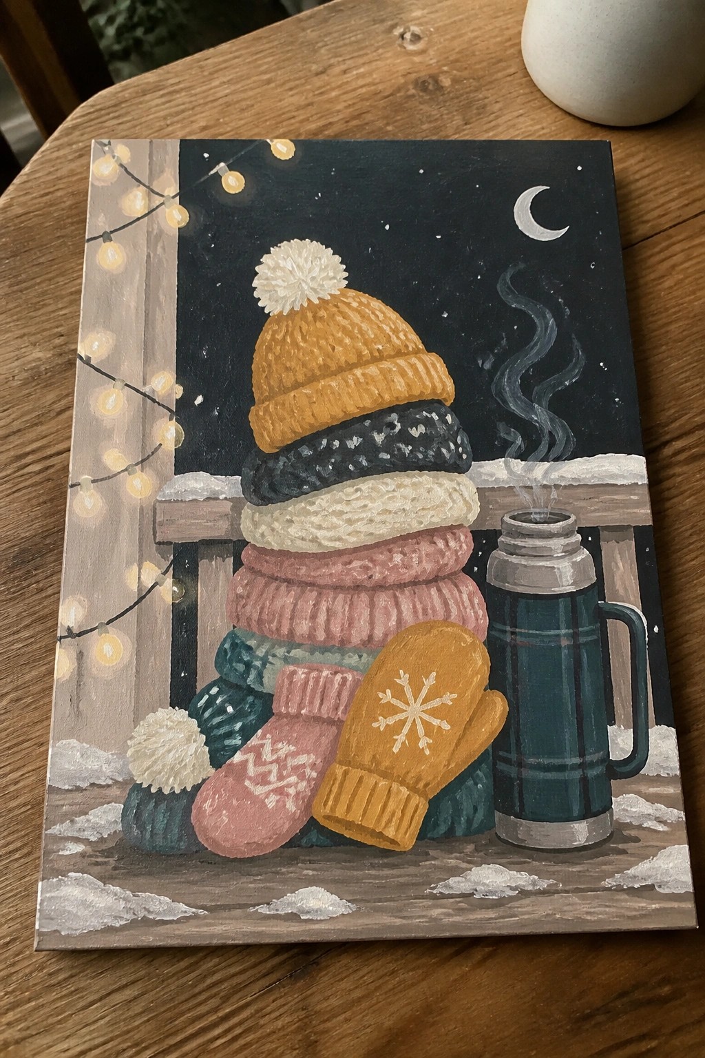 Stacked colorful knit hats, mittens, and steaming thermos on snowy night ledge with lights.
