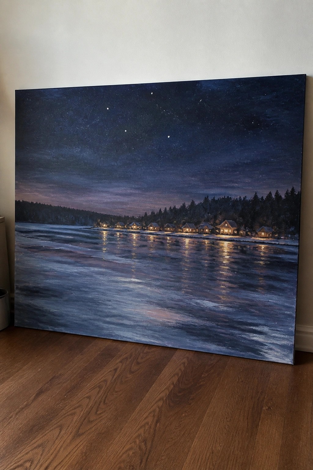 Large painting of starry sky over lit cabins reflected in frozen lake