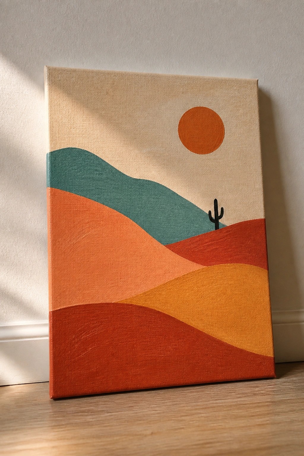 A canvas painting of a desert landscape with layered orange and teal hills, a black cactus silhouette, and an orange sun on a beige background.