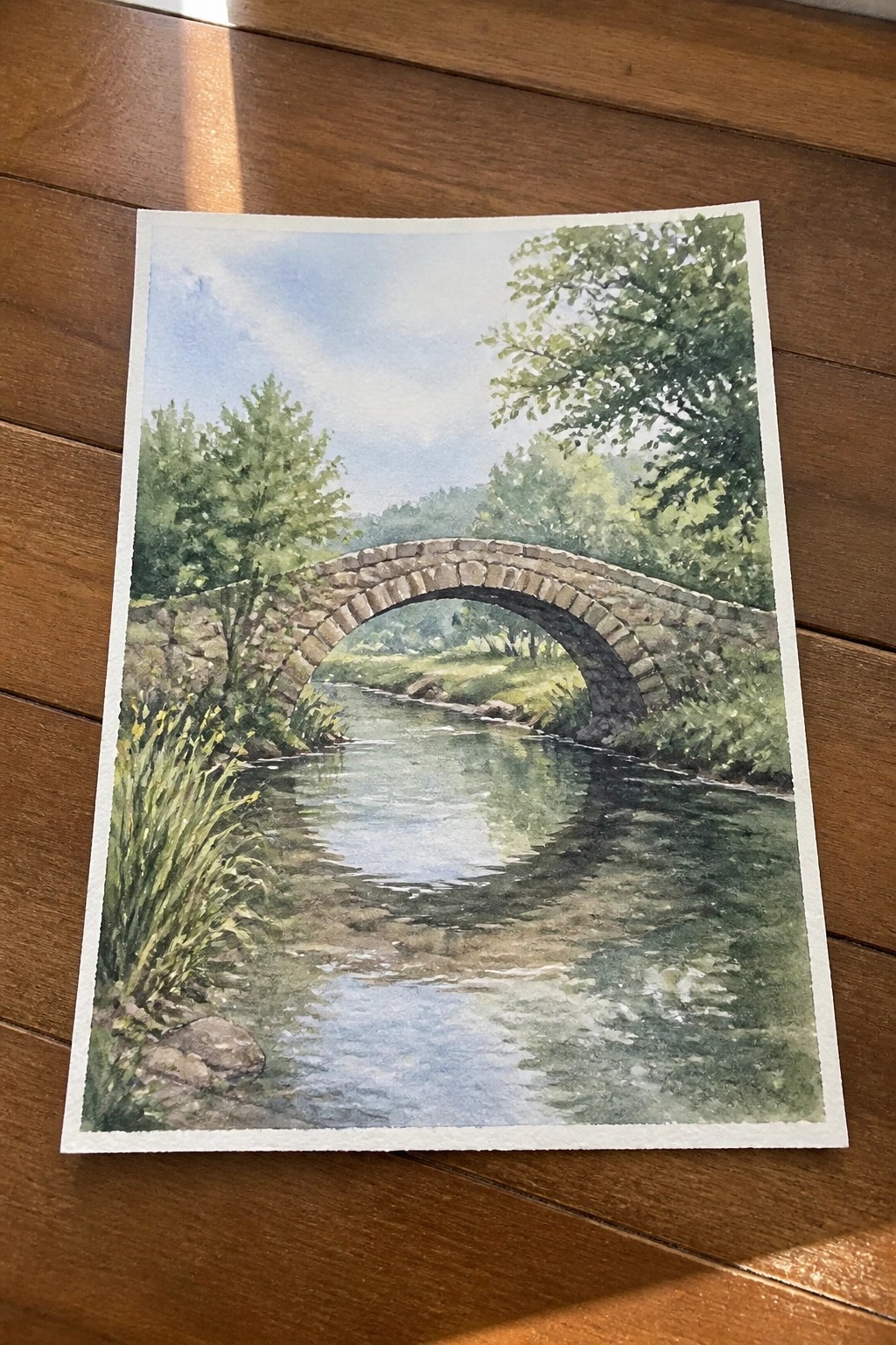 Watercolor painting of stone arch bridge over calm river amid green trees and foliage