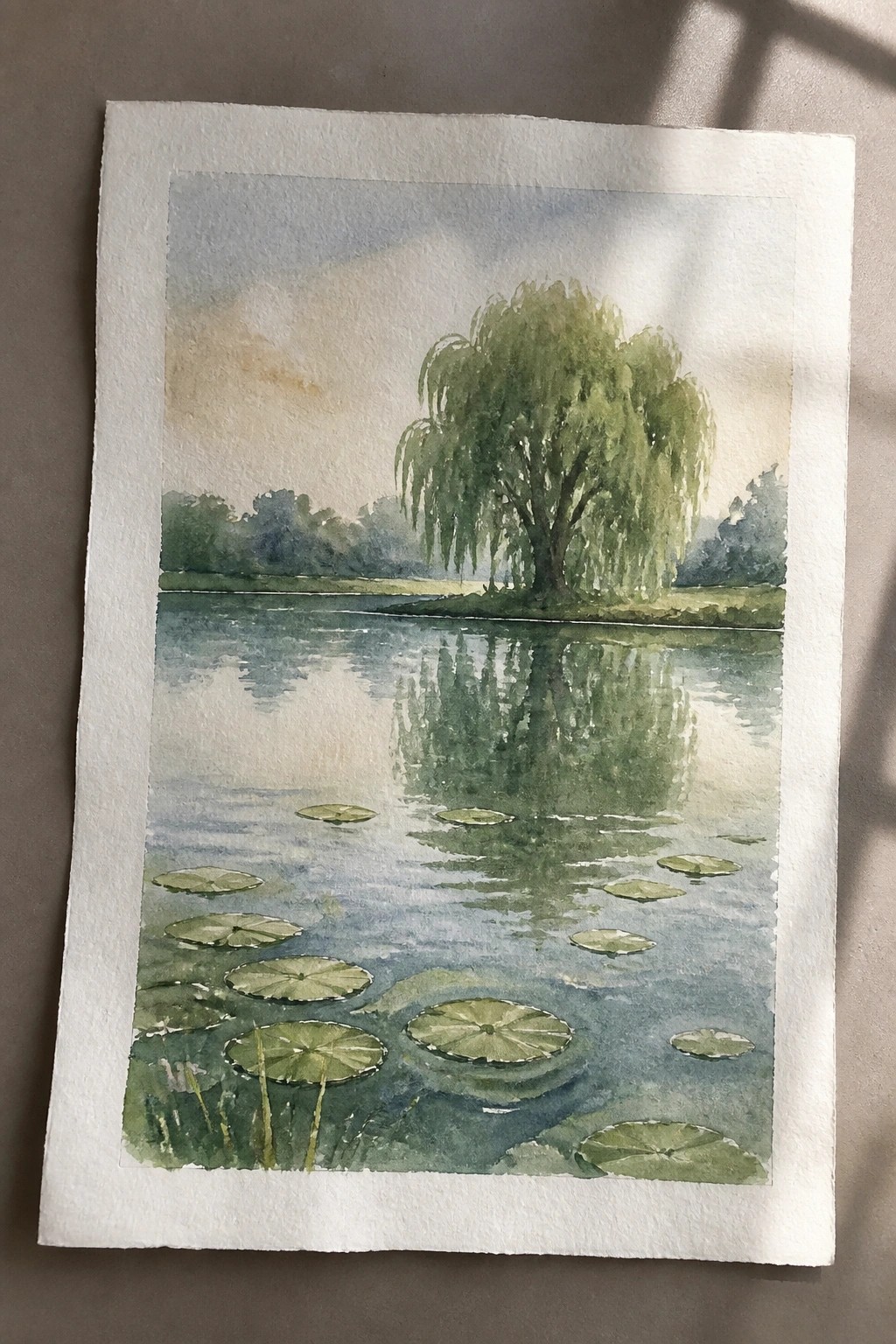 Watercolor of a pond with green lily pads and a weeping willow reflected in calm water.