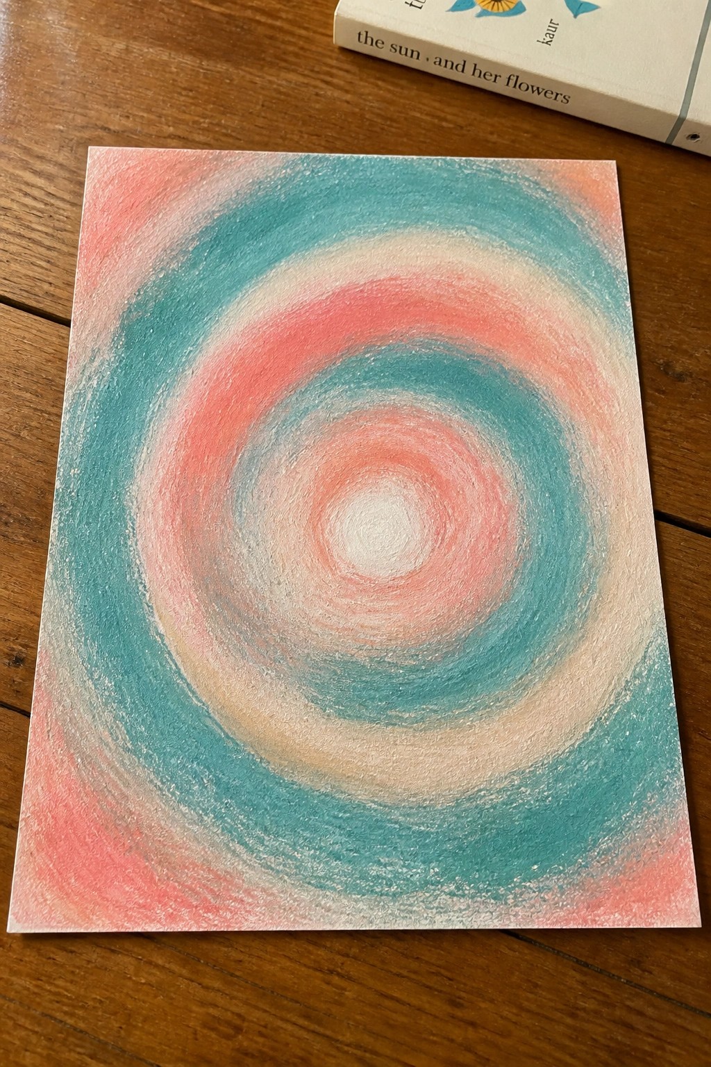 Swirling pink, teal, and white vortex on square paper beside book on wood table