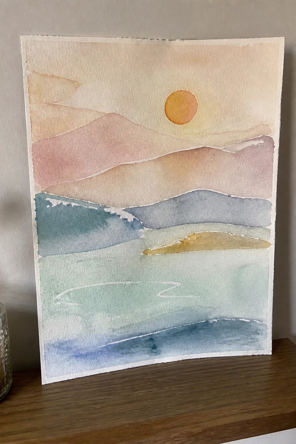 Watercolor landscape with layered pastel hills, orange sun, and blue-green foreground.