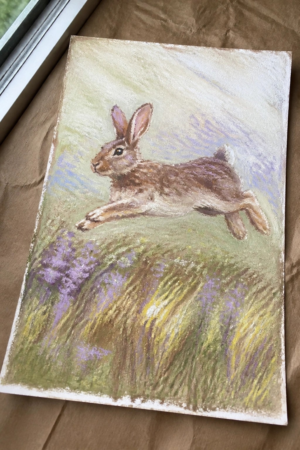 A painting of a brown rabbit leaping across a field of green grass and purple flowers.