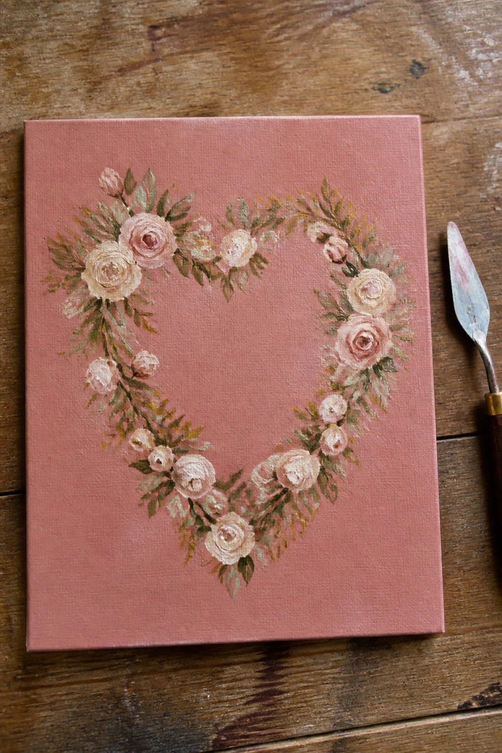Heart-shaped floral wreath in soft pinks on dusty rose canvas beside palette knife.