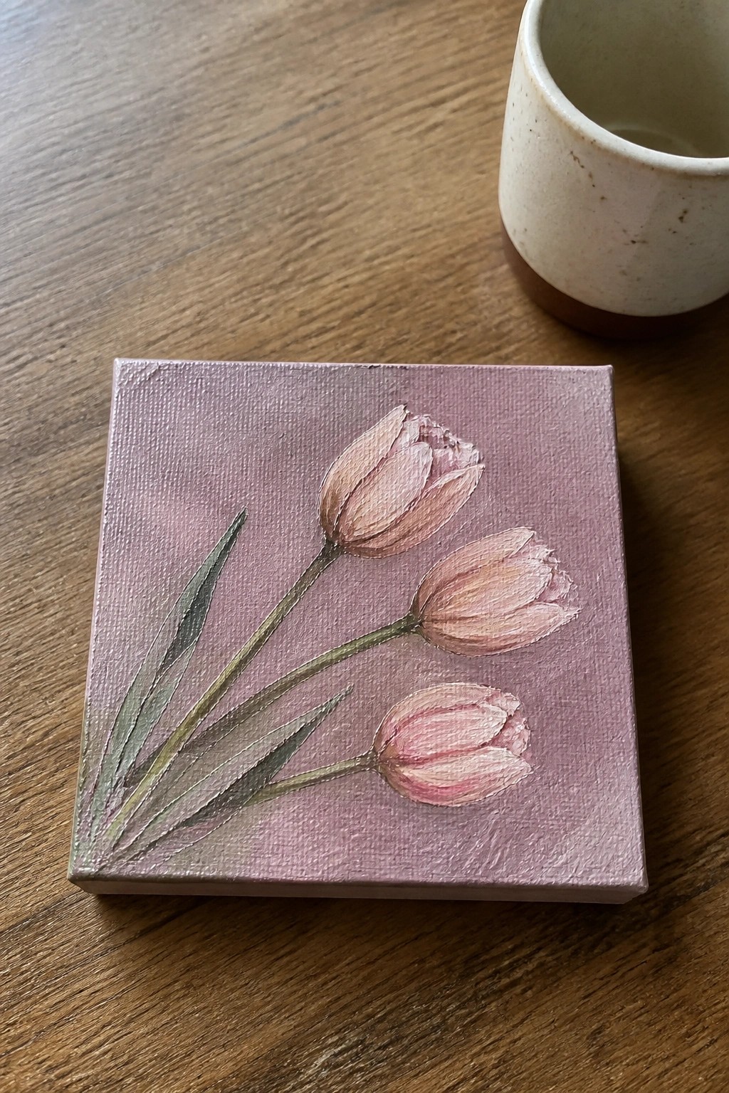 Three pink tulips on small square purple canvas beside white cup on wood.