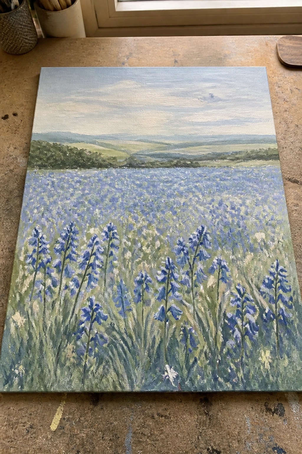 Acrylic painting of a blue wildflower field with green hills in the background on a wooden table.