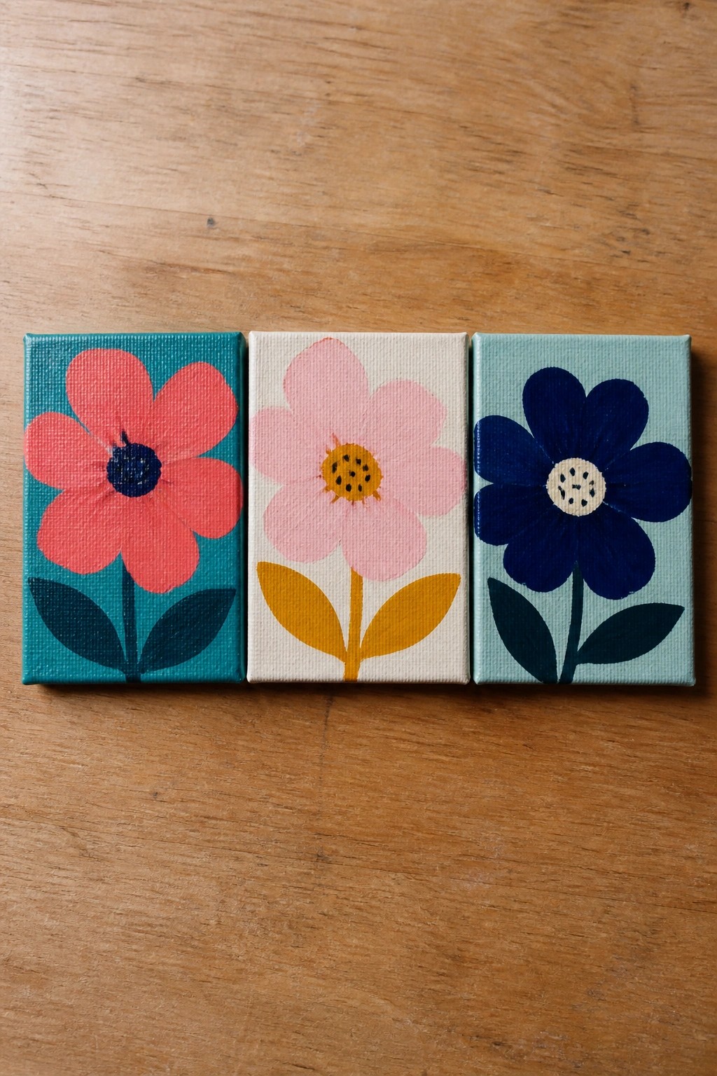 Three colorful flower paintings on small canvases atop a wooden surface.