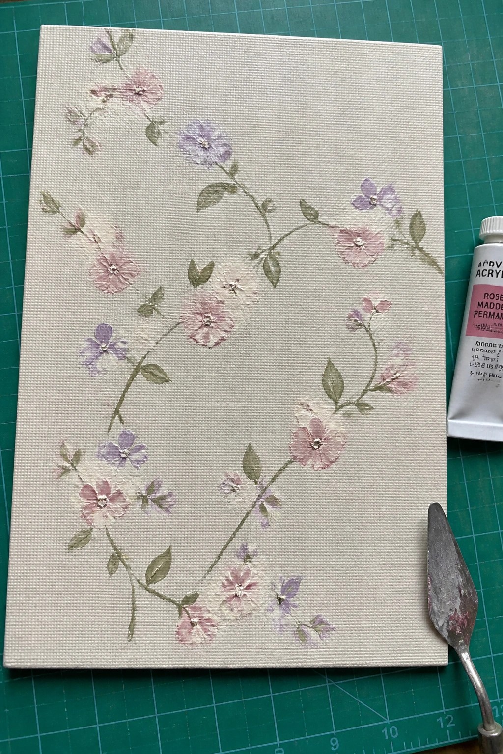 Canvas with pink and lavender flowers, paint tube and knife on green cutting mat.