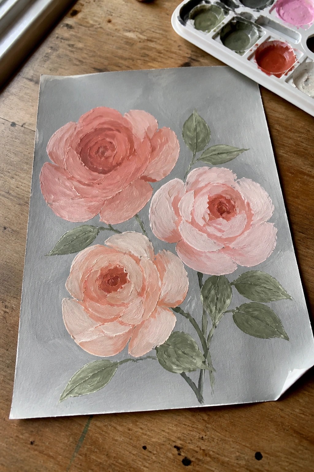 Three pink roses with green leaves painted on gray paper beside a palette