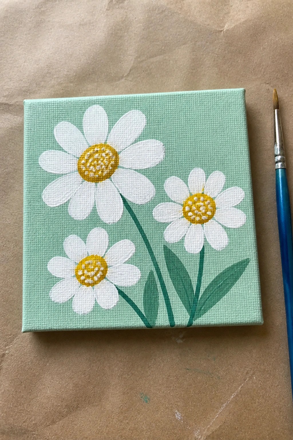 Three daisies with white petals and yellow centers on light green canvas beside paintbrush