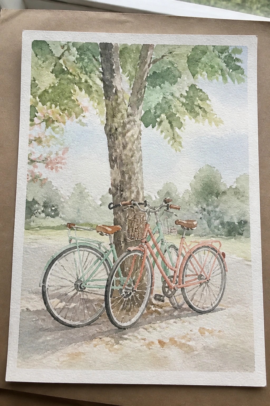 Watercolor painting of pink and mint green bicycles leaning against a tree trunk