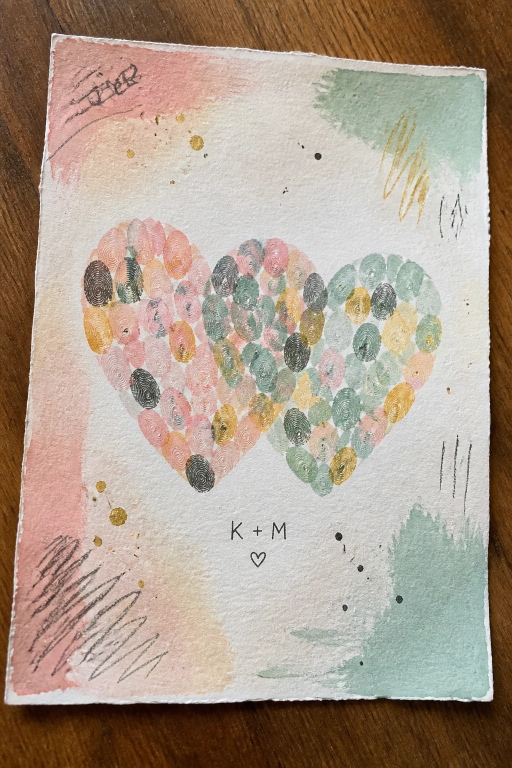 A heart shape made from multicolored fingerprints on a pink and green watercolor background with K + M written below.