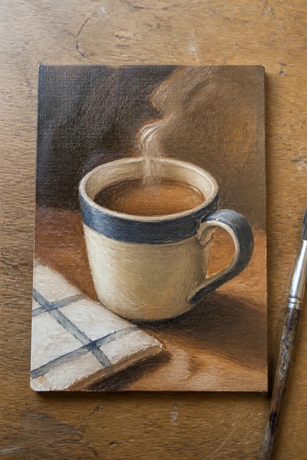 Oil painting of steaming blue-rimmed coffee mug beside checkered cloth and paintbrush on wood