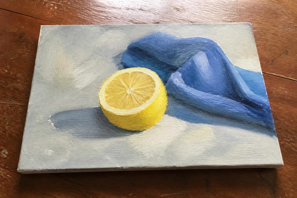 Still life painting of halved yellow lemon beside blue draped cloth on canvas