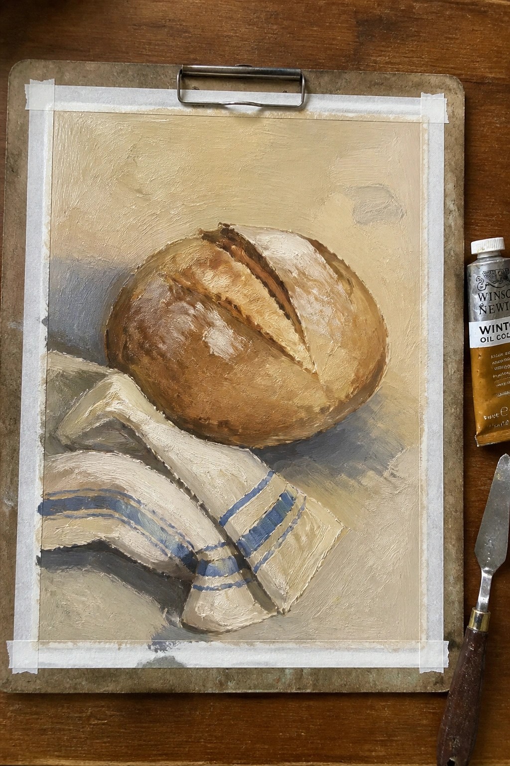 Oil painting of crusty bread loaf on blue-striped cloth beside paint tube and knife