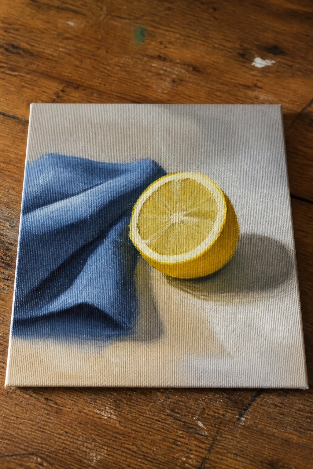 Still life painting of halved yellow lemon beside blue draped cloth on canvas