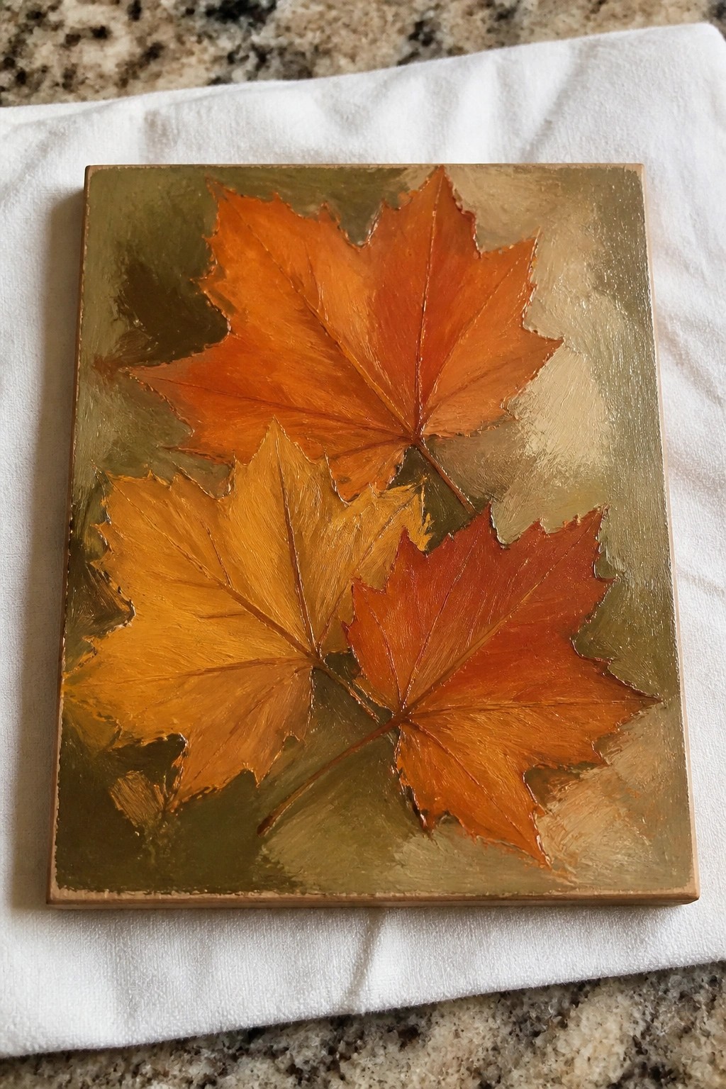 Three autumn maple leaves in orange tones on gold square canvas over white cloth.