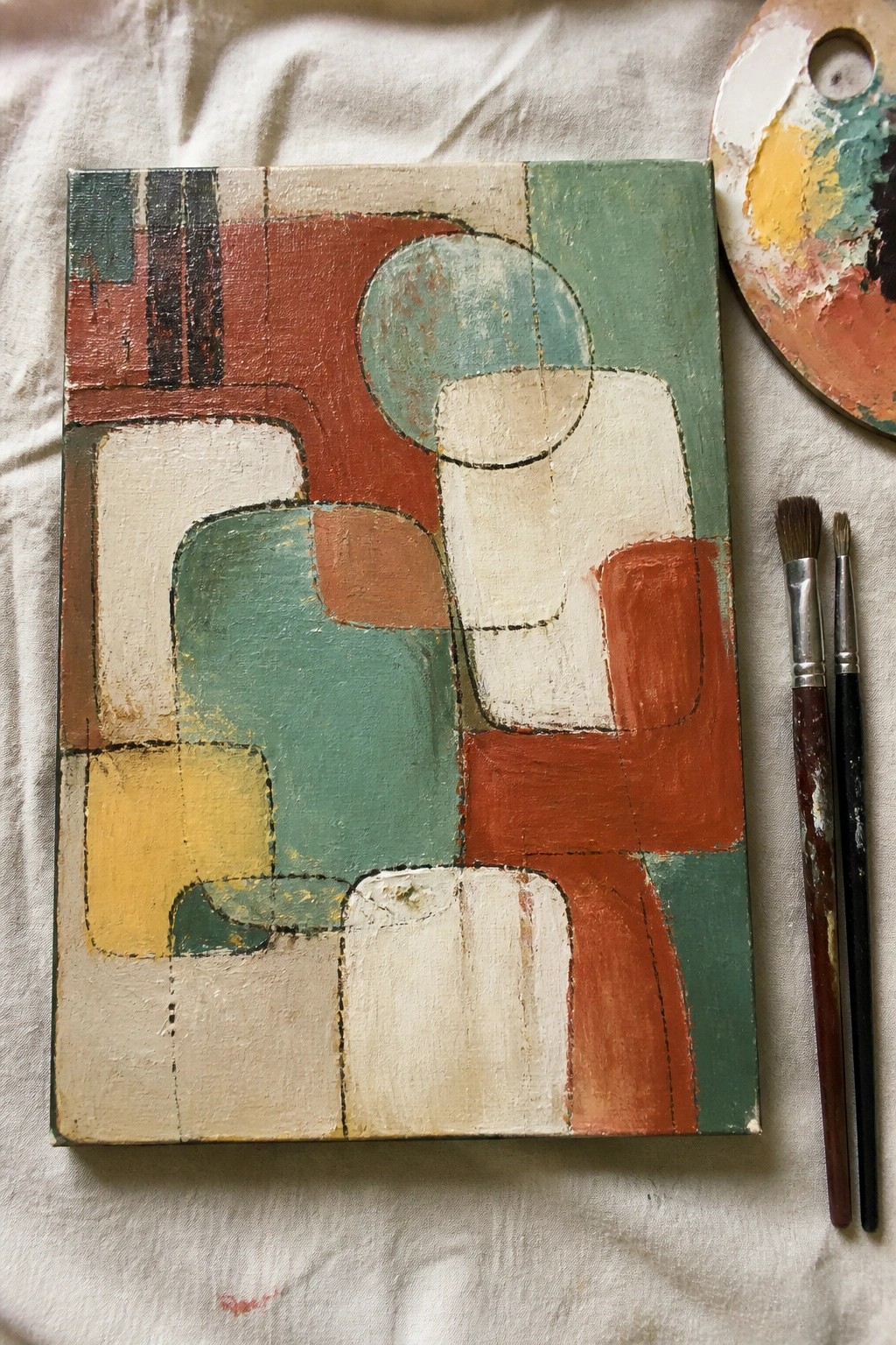 Abstract painting with geometric shapes in red, teal, and cream beside palette and brushes
