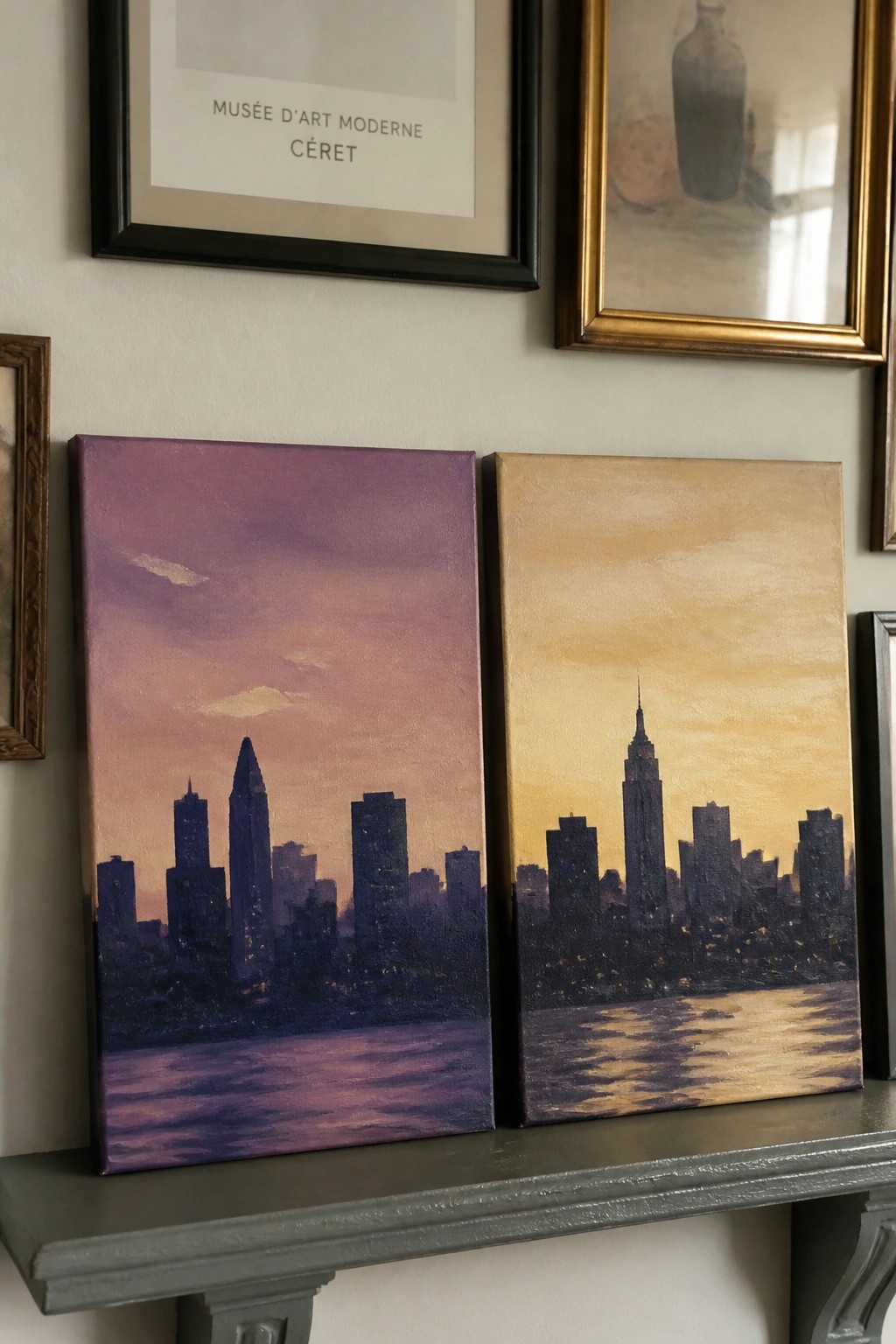Two city skyline paintings with purple and gold skies on a gray shelf.
