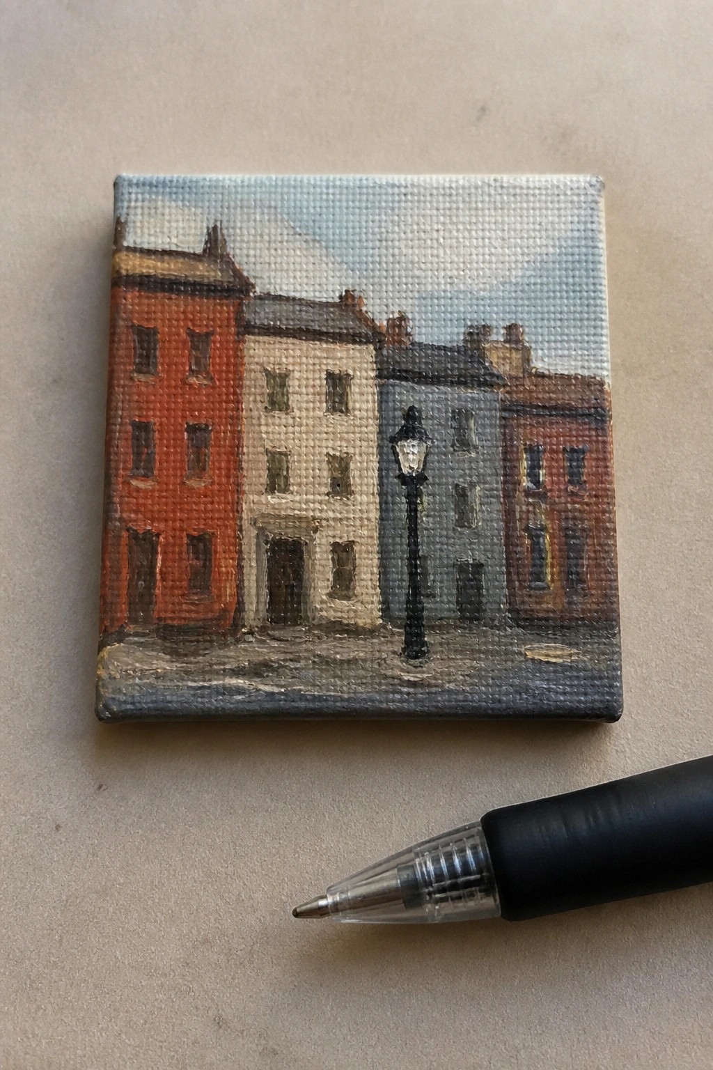 Miniature canvas painting of colorful buildings with lamppost beside a black pen for scale.