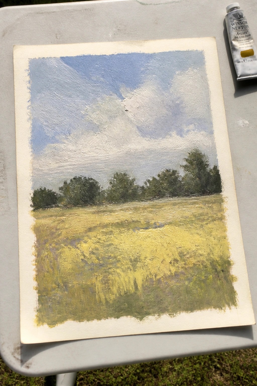 Oil painting of yellow field, green trees, and blue cloudy sky beside yellow ochre tube