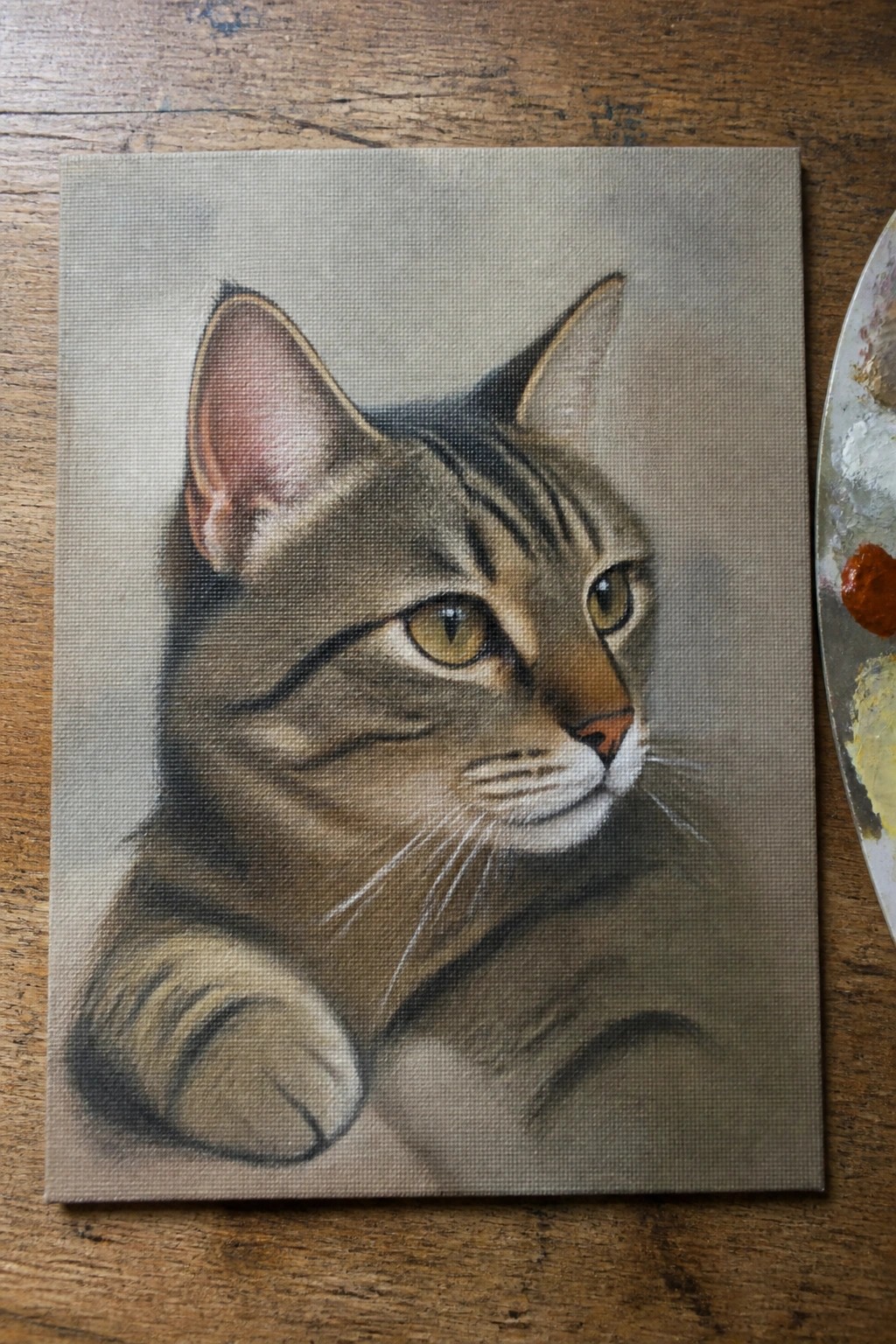 A realistic oil painting of a brown tabby cat's face and paw resting on a canvas next to a paint palette on a wooden surface.