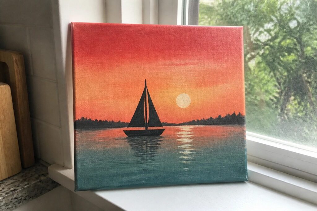 22 Practical Canvas Painting Ideas Designed for Paint-and-Sip Nights A canvas painting of a red-to-orange sunset over teal water with a black sailboat silhouette and sun reflection, resting on a windowsill.