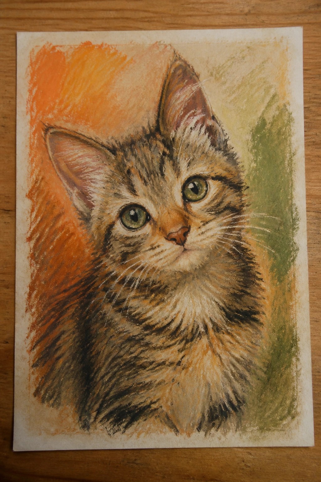 Pastel drawing of fluffy tabby kitten with green eyes on paper over wood