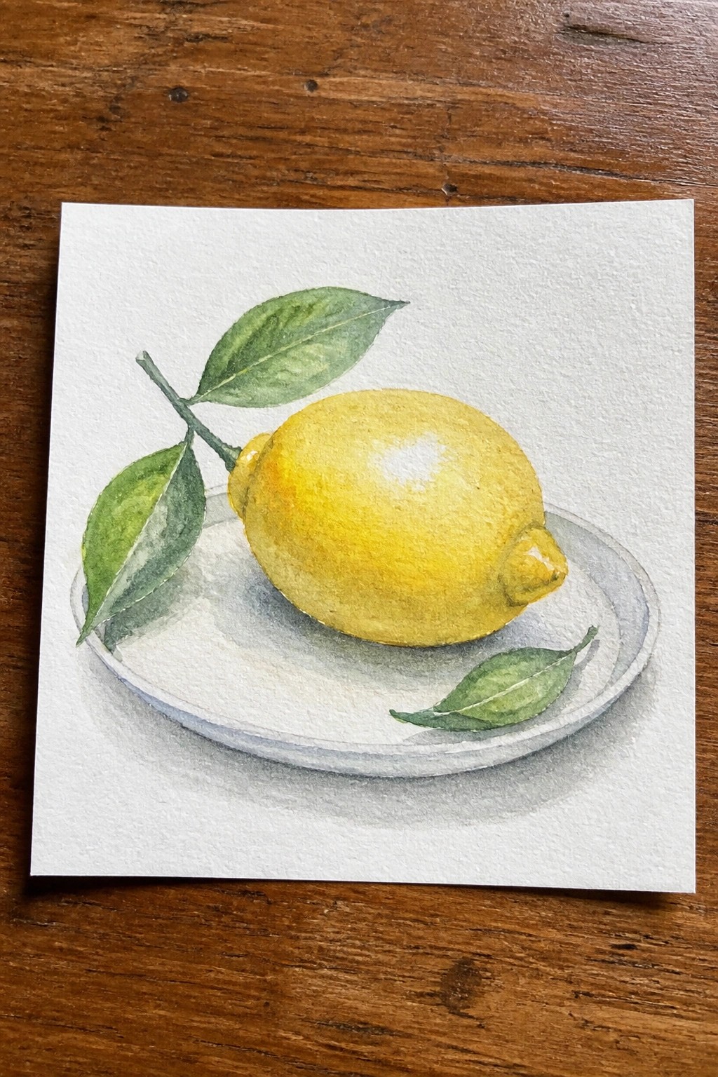 Watercolor of a yellow lemon with green leaves on a white plate atop wood.