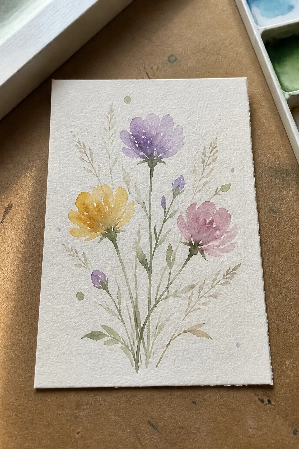A watercolor painting of yellow, purple, and pink flowers with green stems and leaves on white paper.