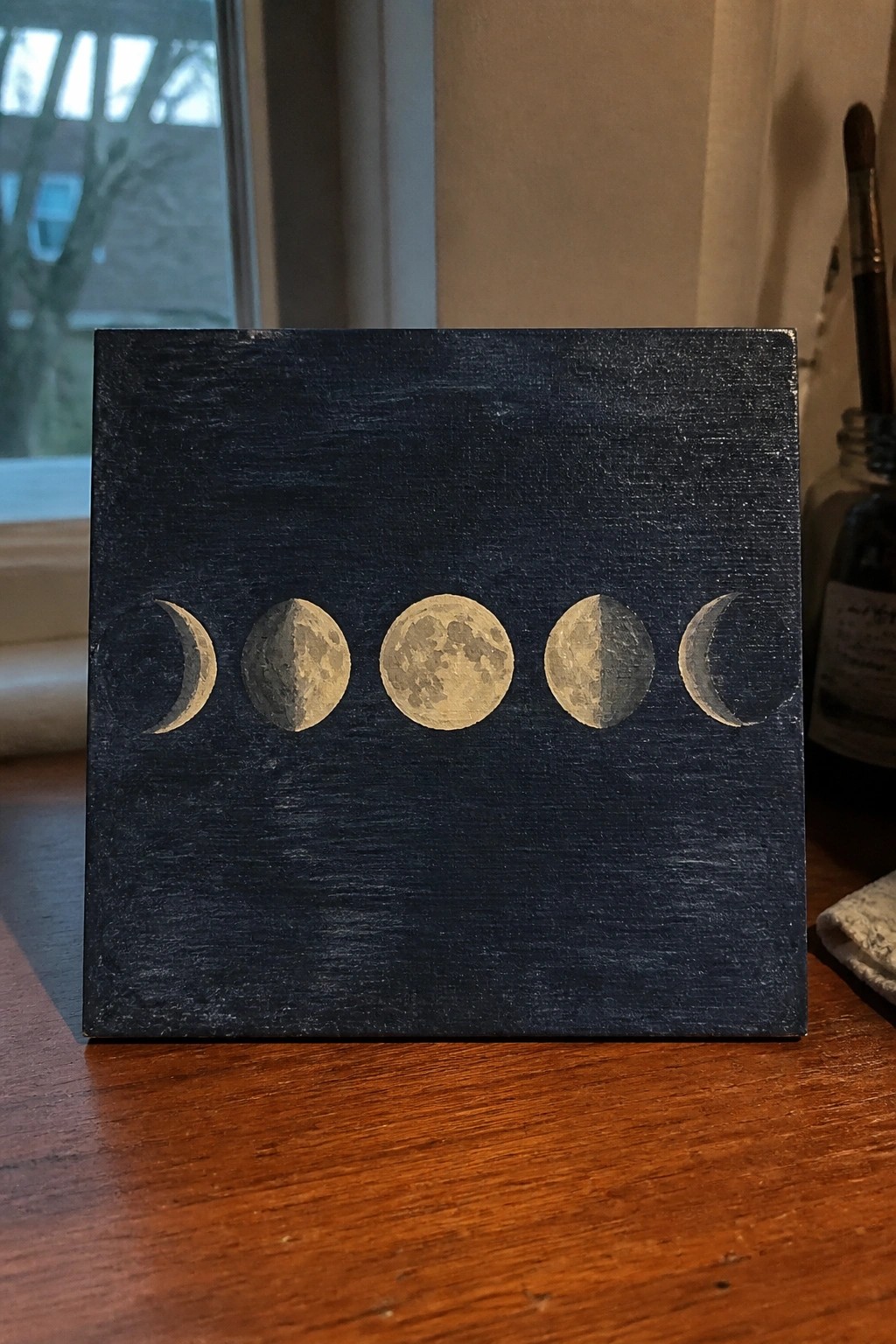 Dark blue square canvas with five golden moon phases painted in a row on wood.
