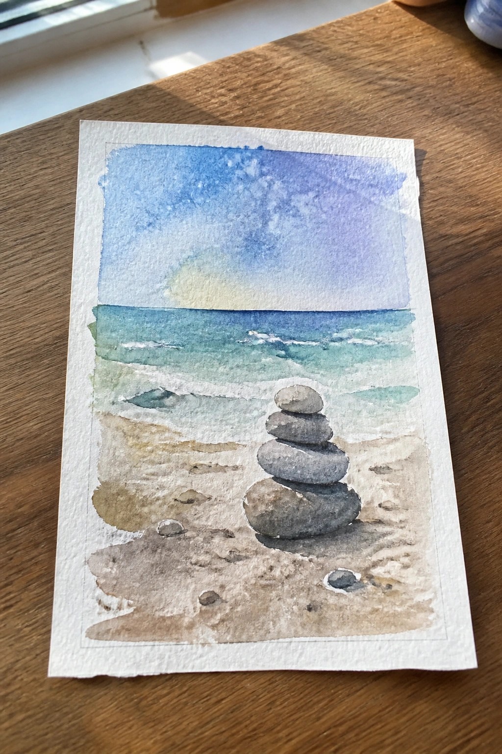 Watercolor painting of stacked stones on beach with ocean waves under sunset sky.