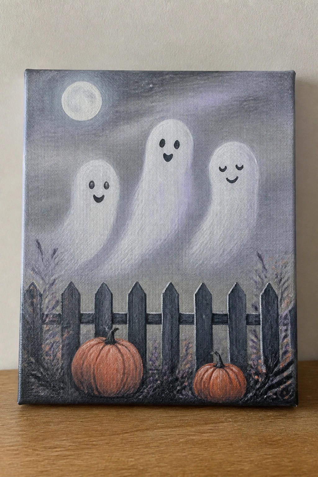 Three white ghosts float behind a dark fence with orange pumpkins under a full moon