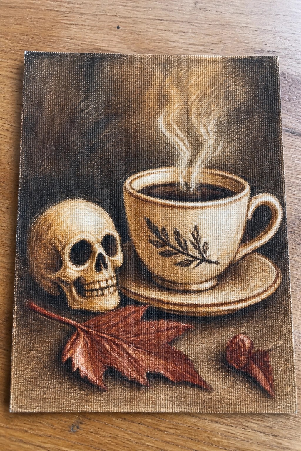 Human skull beside steaming coffee cup, red maple leaf, and nut on canvas.