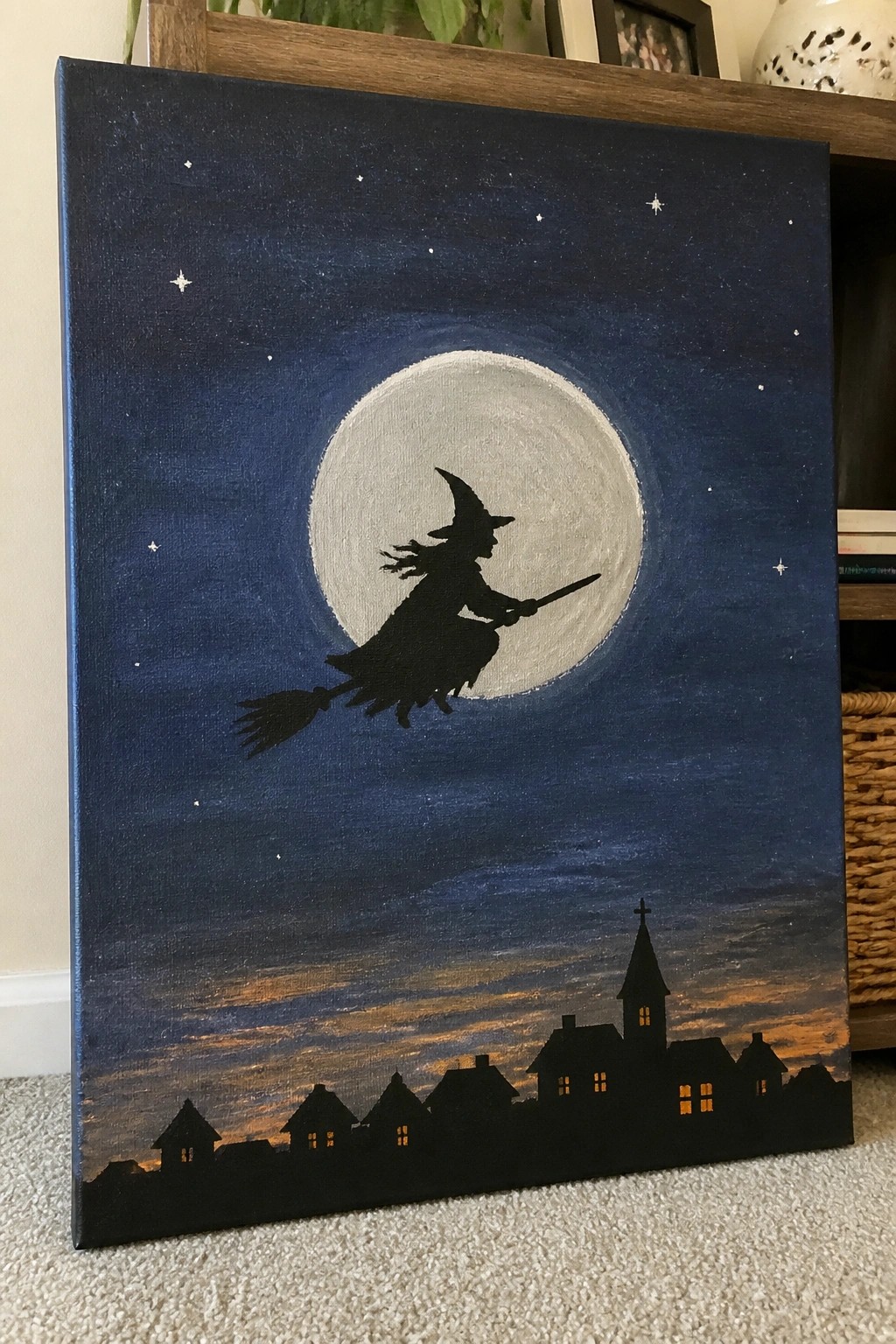 Witch silhouette on broomstick flies before full moon above silhouetted village at night