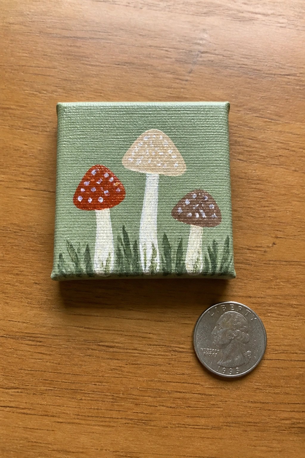 Small canvas with three dotted mushrooms beside a 1998 US quarter on wood