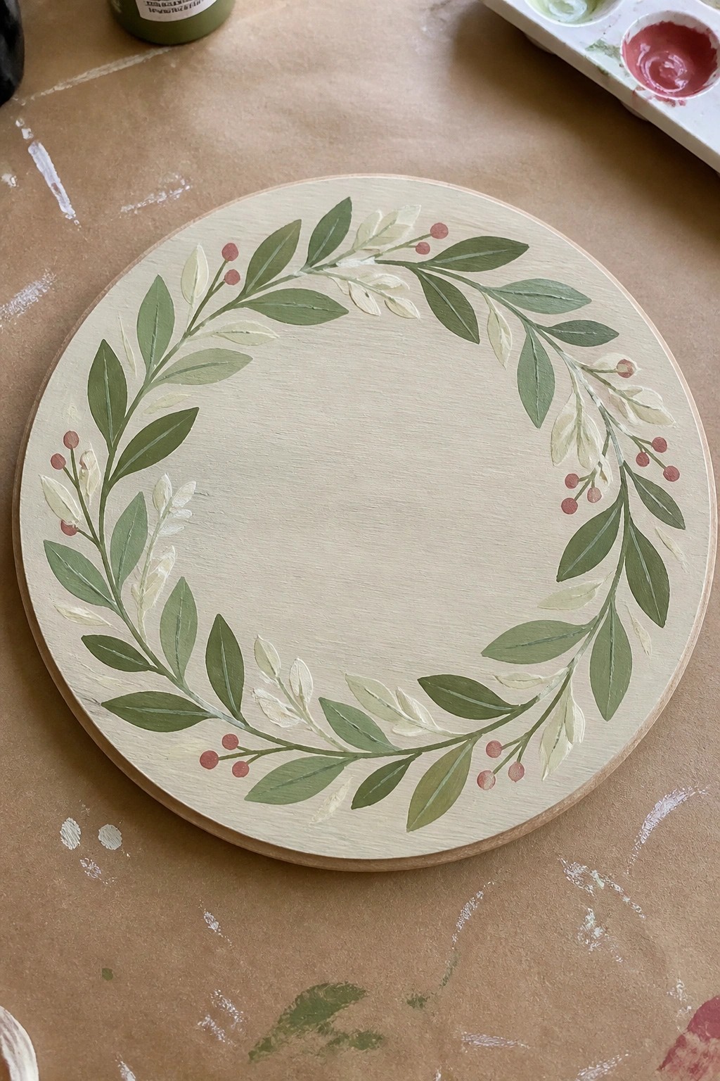 Round wooden plaque with hand-painted green leaf wreath and red berries on craft table.
