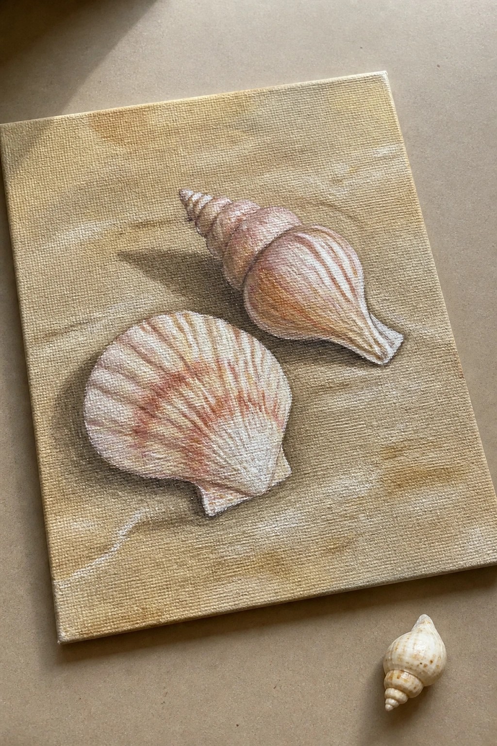Canvas painting of spiral and scallop seashells beside small real shell on beige surface.