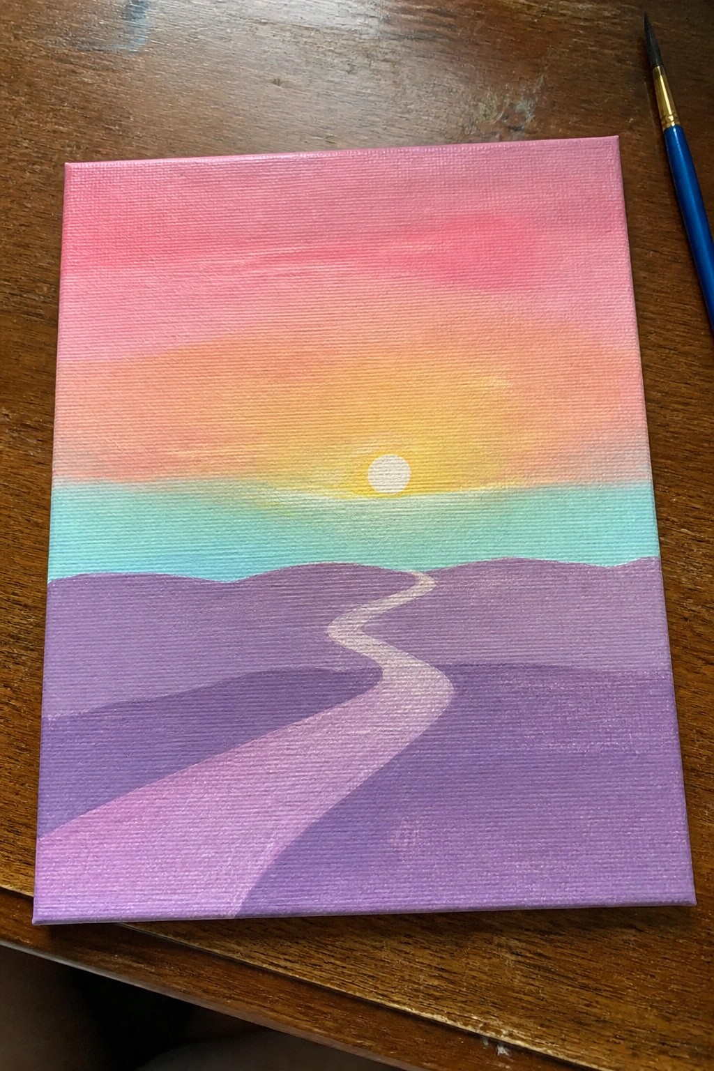 Sunset painting with pink to yellow sky, sun over turquoise, purple hills with white path.