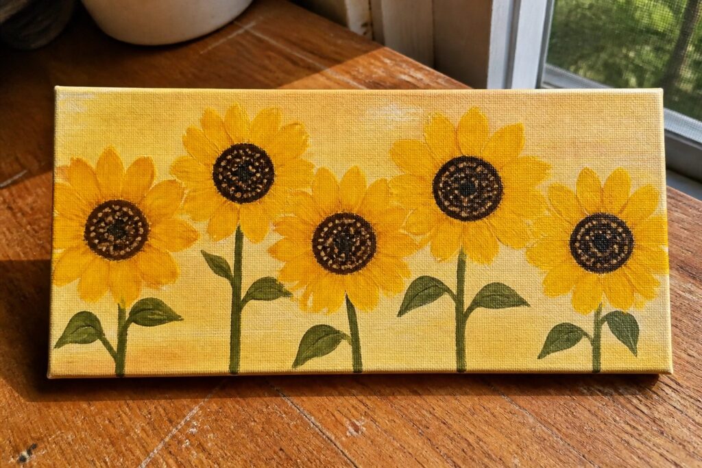 Yellow canvas painting of five sunflowers with green stems on wooden table