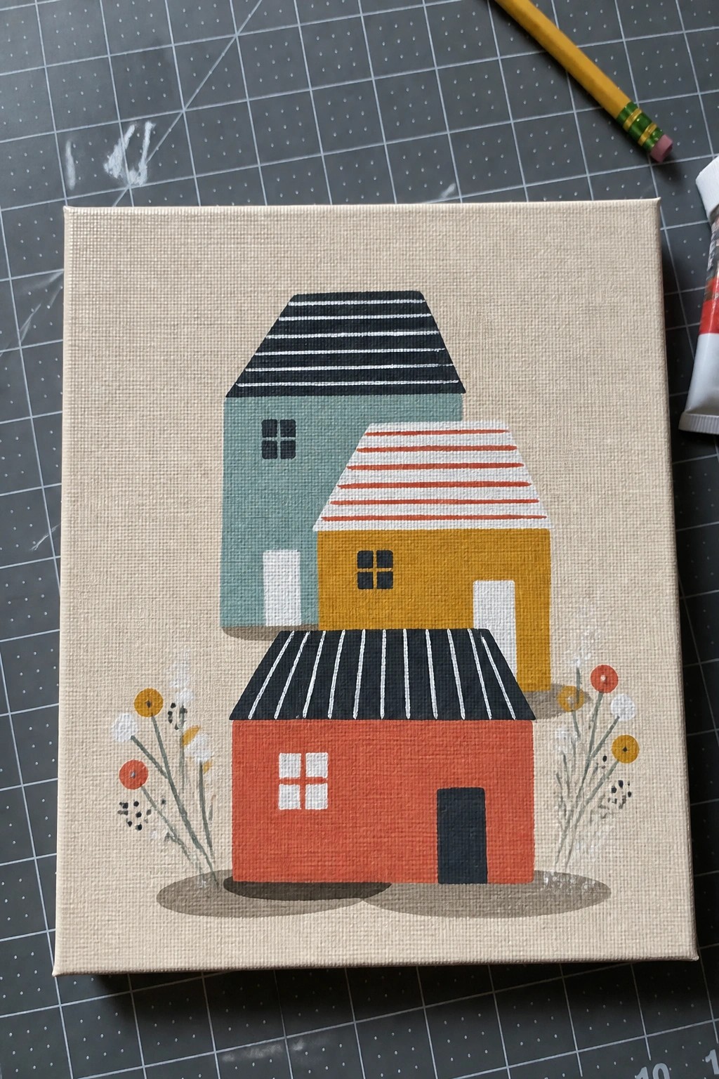 A canvas painting shows three overlapping houses in red, yellow, and teal with striped roofs and flowers at the bottom.