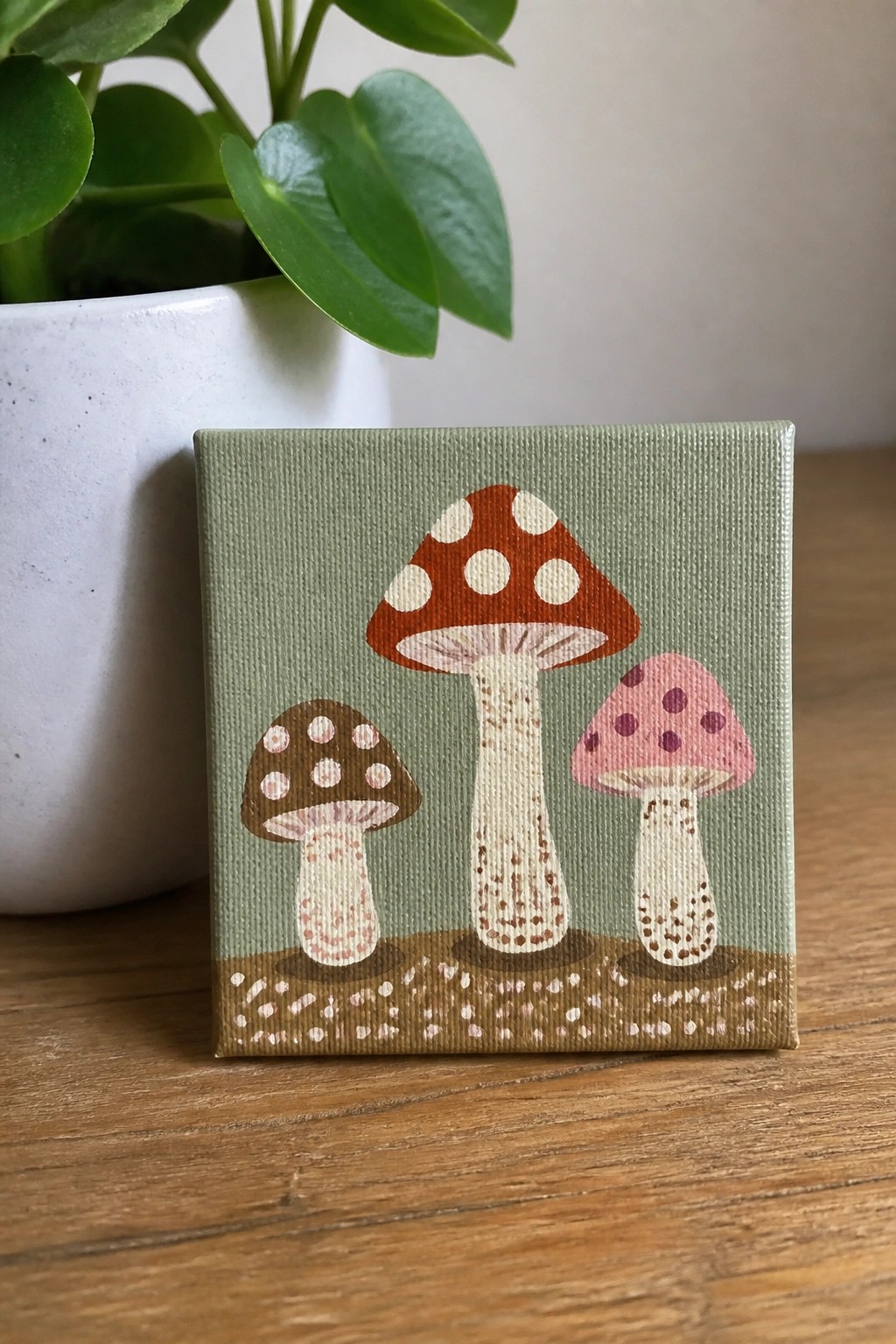 Three mushrooms painted on small green canvas with brown soil and plant nearby.
