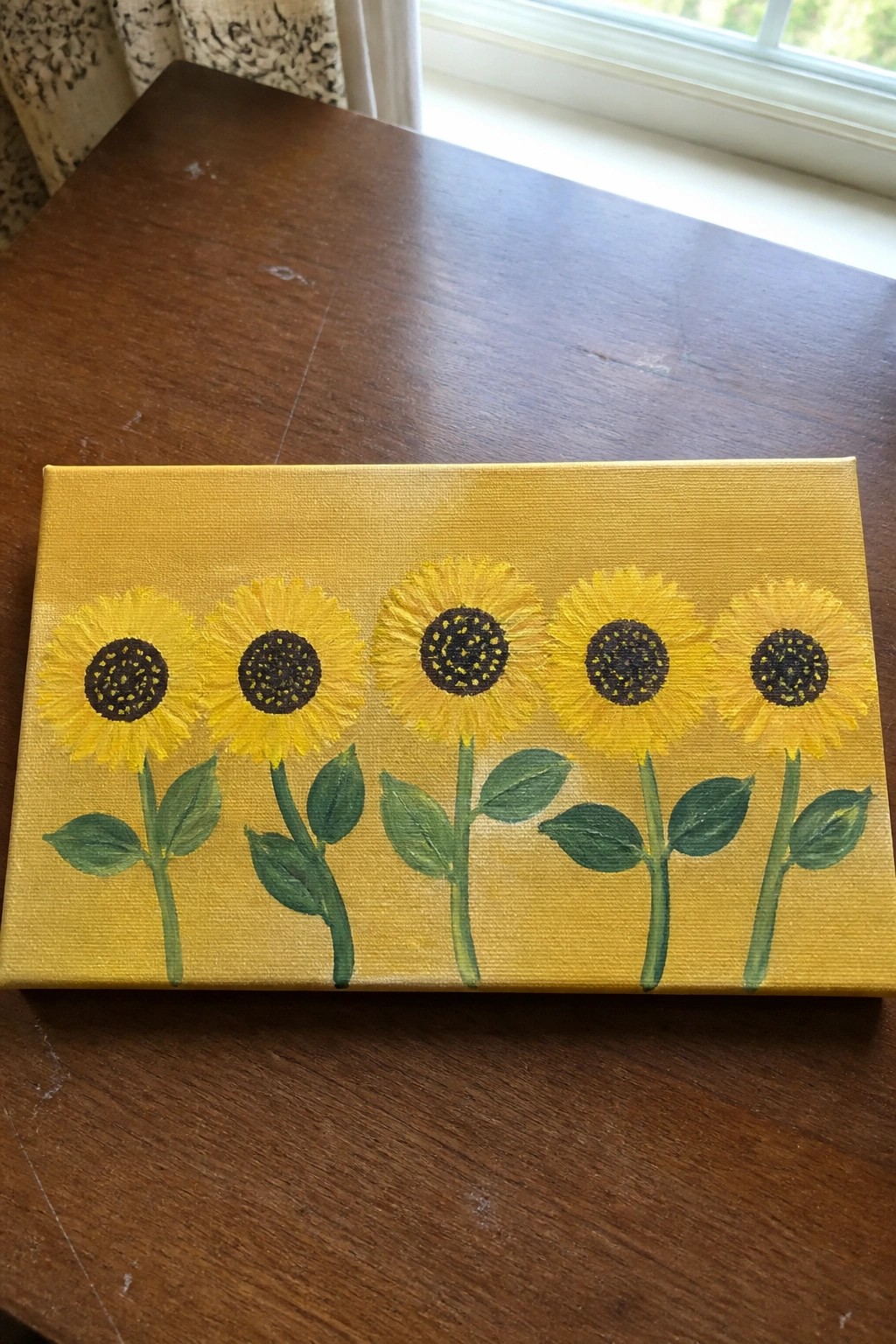 Yellow canvas painting of five sunflowers with green stems on wooden table