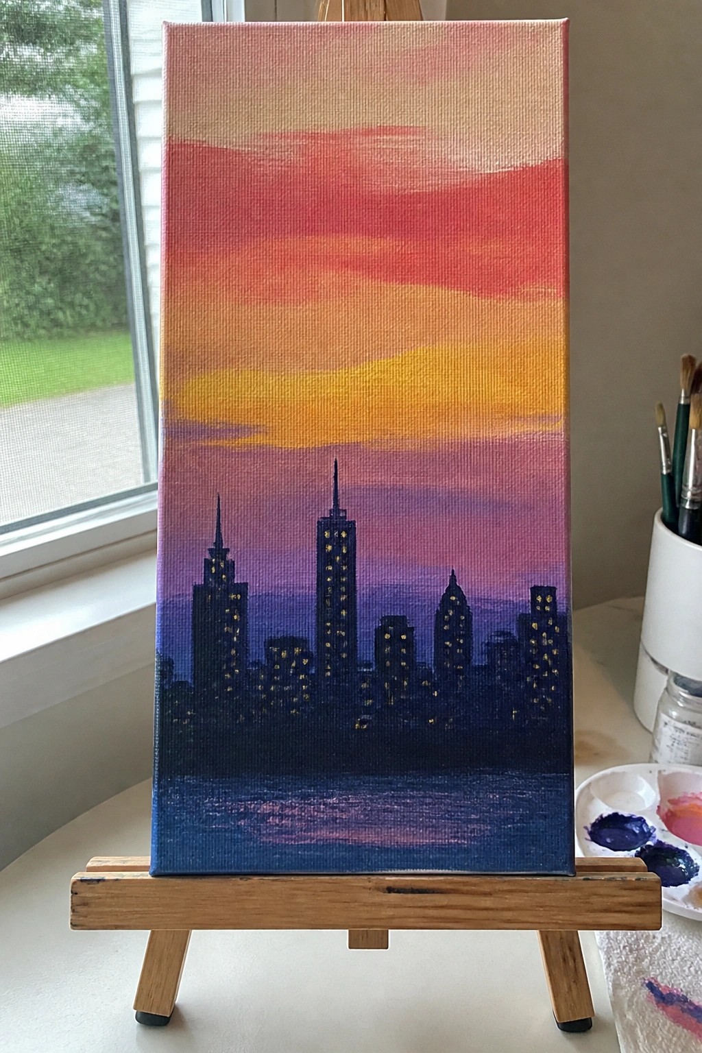 Canvas on easel shows dark city skyline with glowing windows under pink-orange sunset sky.