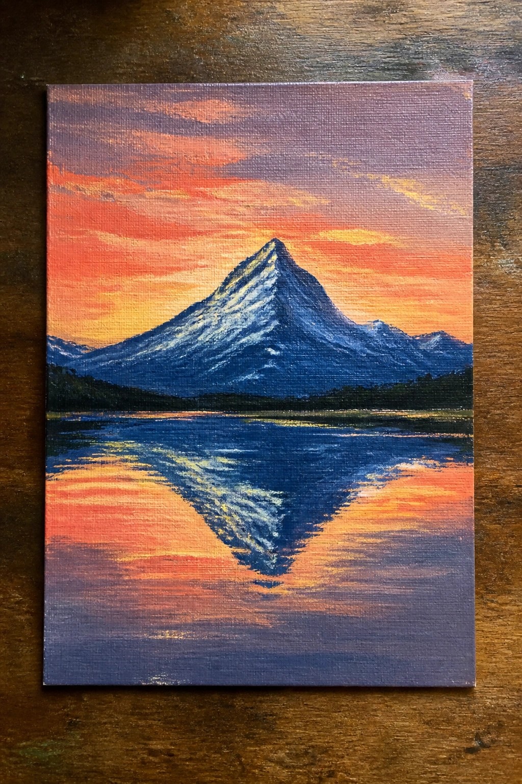 Vibrant sunset painting of snow-capped mountain reflected in colorful lake with orange-purple sky.